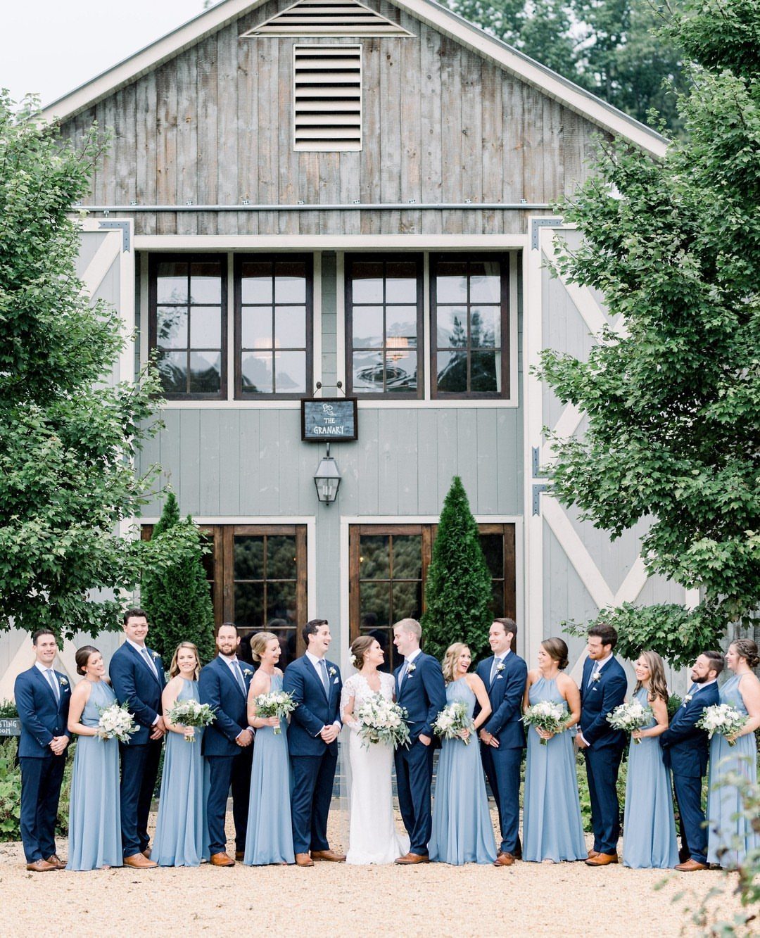 10 Blue Color Palette Wedding Ideas for Every Season 6 Wedding party in navy suits and dusty blue dresses posing before a large grey barn.