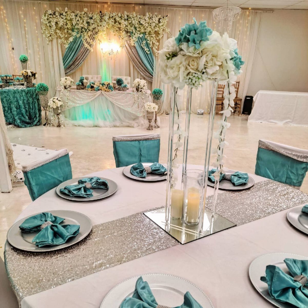 10 Blue Color Palette Wedding Ideas for Every Season 7 Wedding reception tables set with teal napkins, silver sequin runners, and tall floral centerpieces.