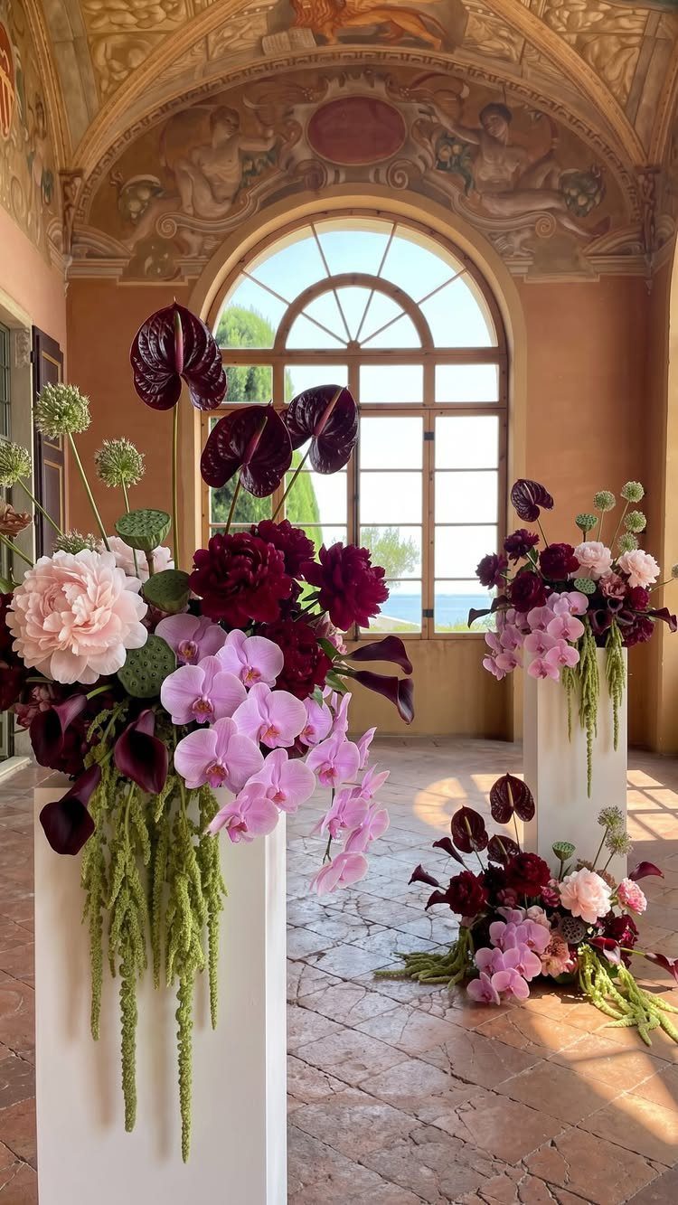 10 Burgundy Wedding Theme Ideas for Tables, Cakes and Attire 2 Burgundy and pink floral arrangements with trailing greenery on white pedestals in a grand room.