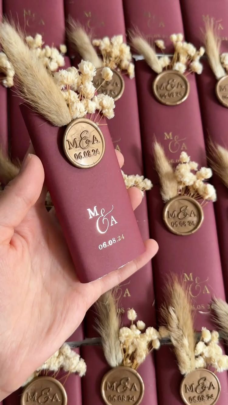 10 Burgundy Wedding Theme Ideas for Tables, Cakes and Attire 8 A hand holding a burgundy wedding favor tube decorated with dried flowers and a gold wax seal.