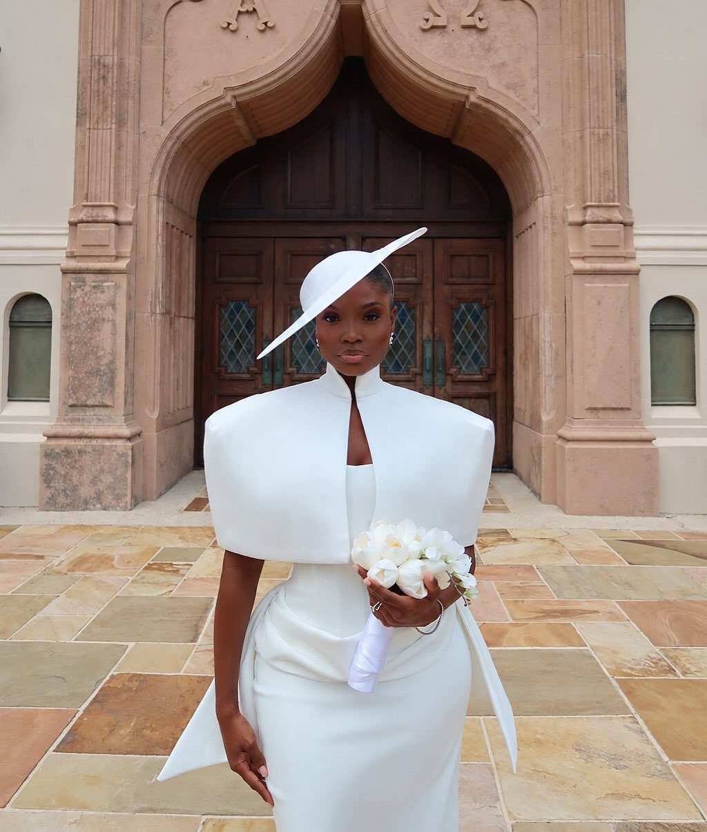 Woman wearing a structured white bridal capelet and wide-brimmed hat holding white flowers.