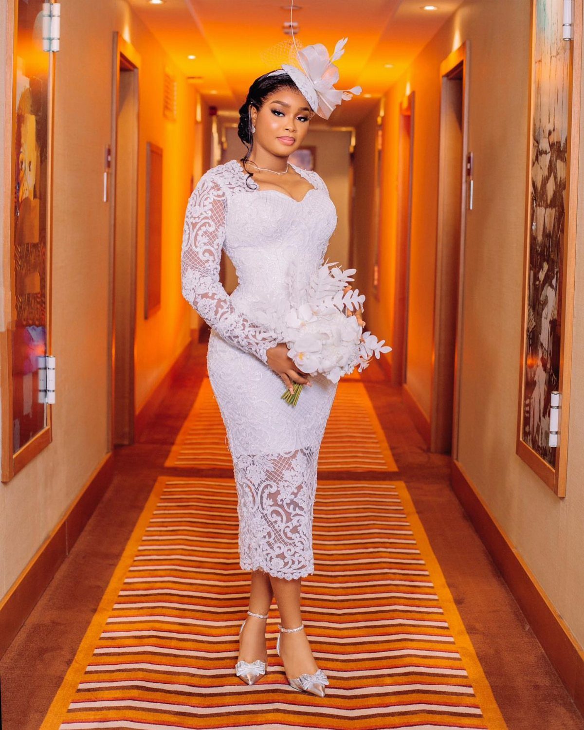 Woman in a white lace tea-length dress posing with a bouquet in a hallway.