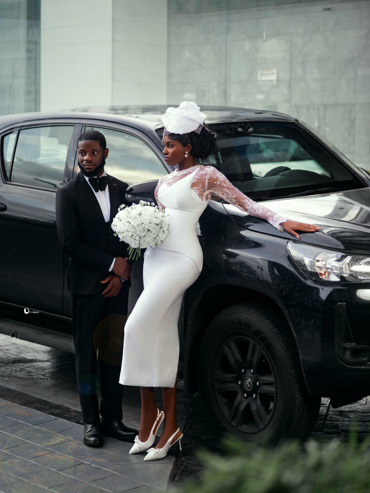 Woman in a white lace sheath dress posing with a bouquet next to a black vehicle.
