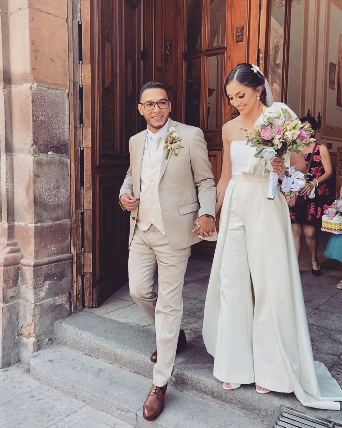 Bride in a white strapless jumpsuit with a long train walking with her groom.