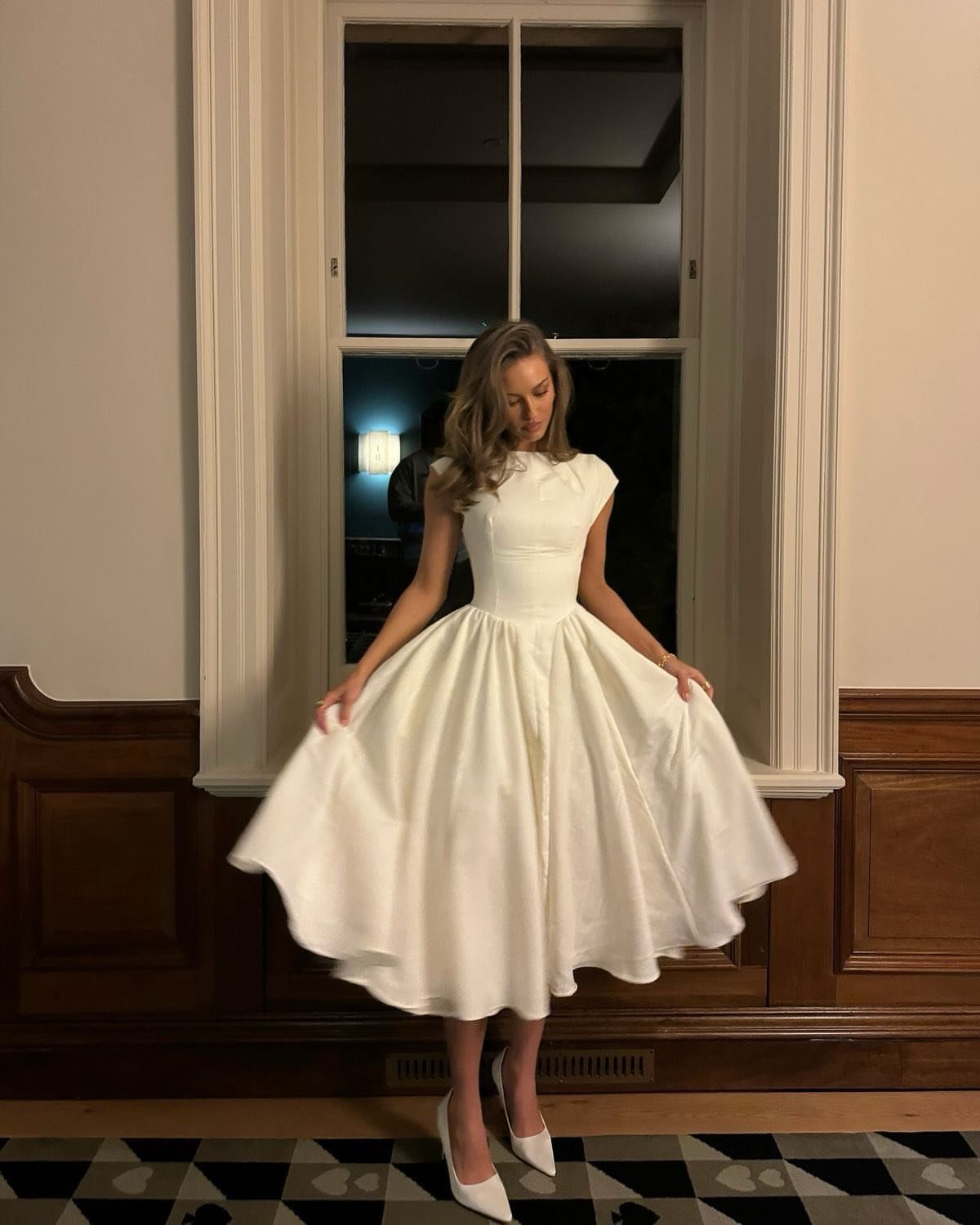 Woman in a white sleeveless dress with a full pleated skirt standing before a window.