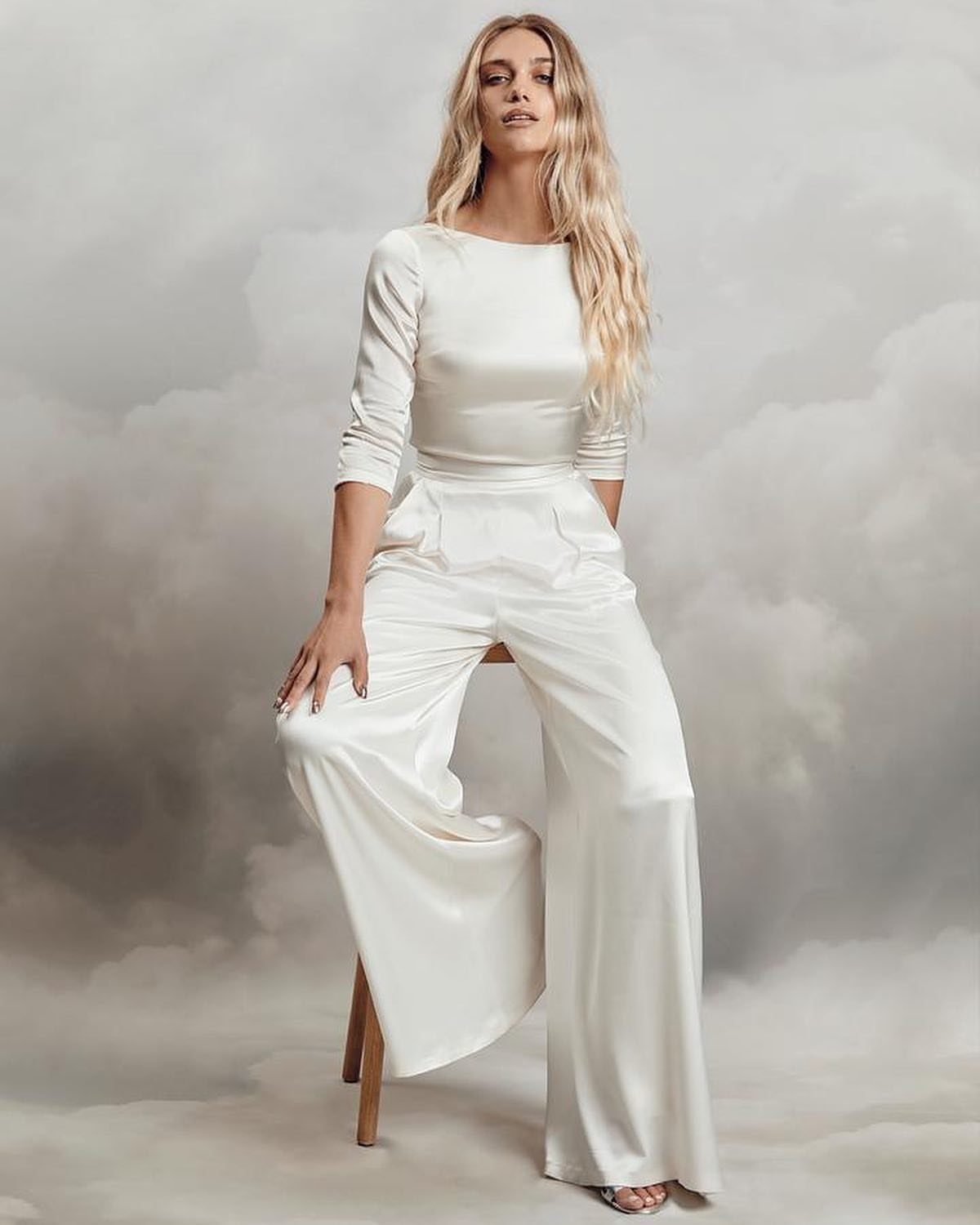 A blonde woman wearing a white satin long-sleeved top and wide-leg trousers sitting against clouds.