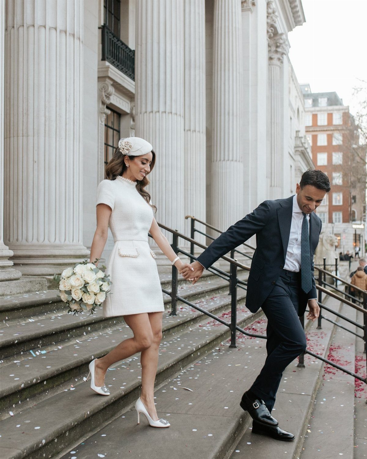 A bride in a white mini dress and a groom in a navy suit walk down stone steps.