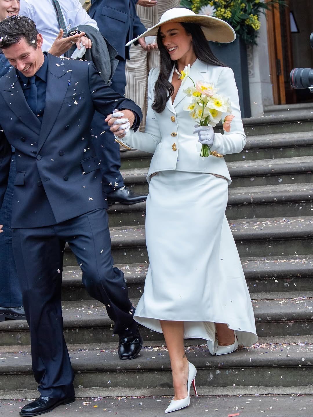 A woman in a white skirt suit and large hat walks down stairs holding yellow flowers.