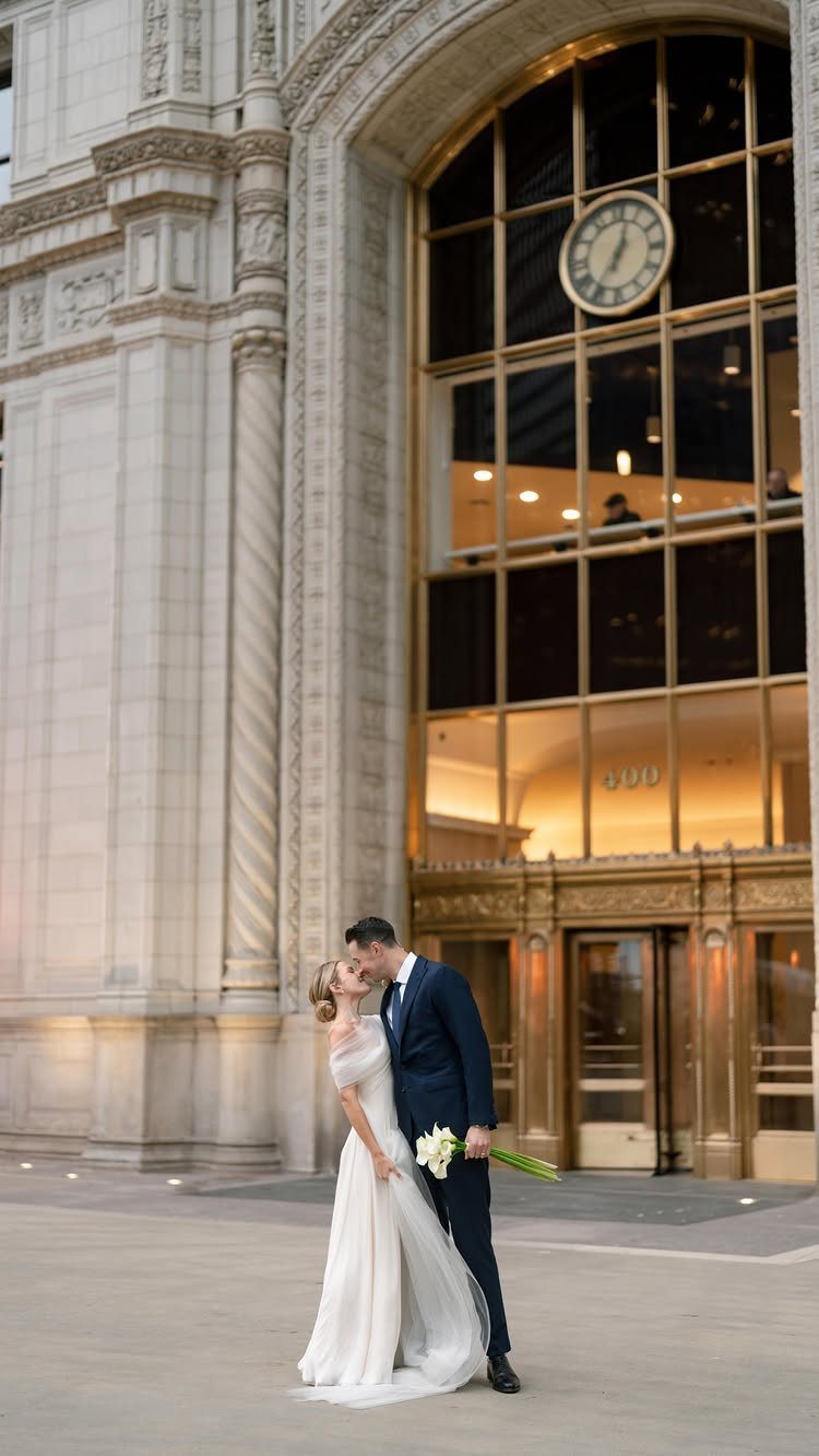 A bride in a white A-line dress and a groom kissing in front of a grand building.