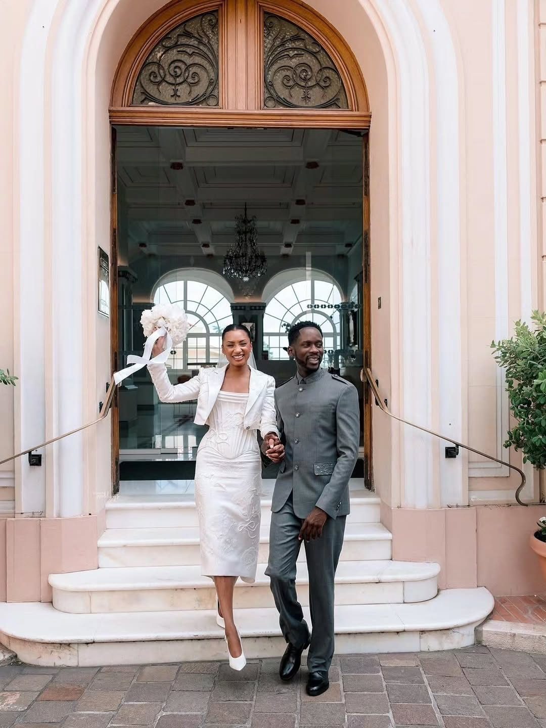 A bride in a white midi wedding dress and jacket walks down steps with her groom.