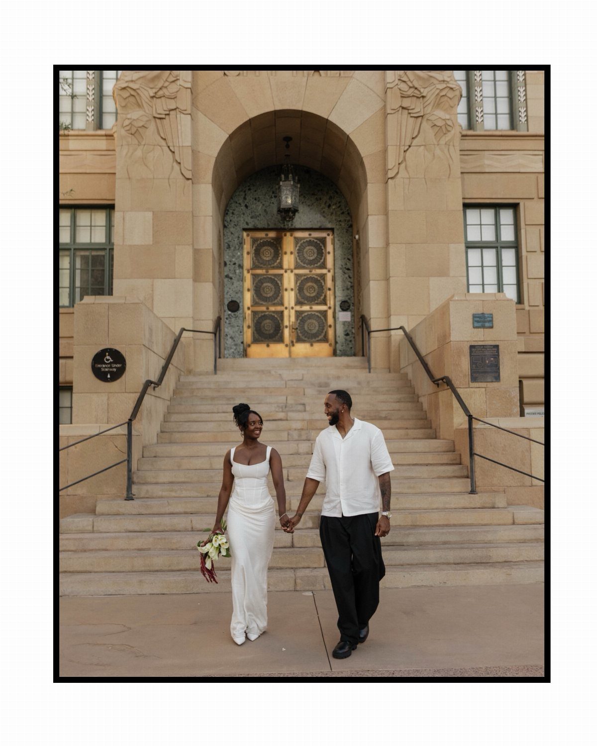 A bride in a minimalist white dress walks down stone steps holding hands with her groom.