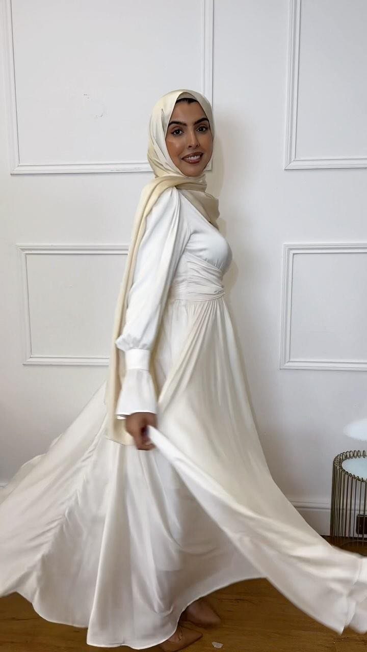 Woman in a long-sleeved white satin wedding dress and cream hijab posing against a white wall.
