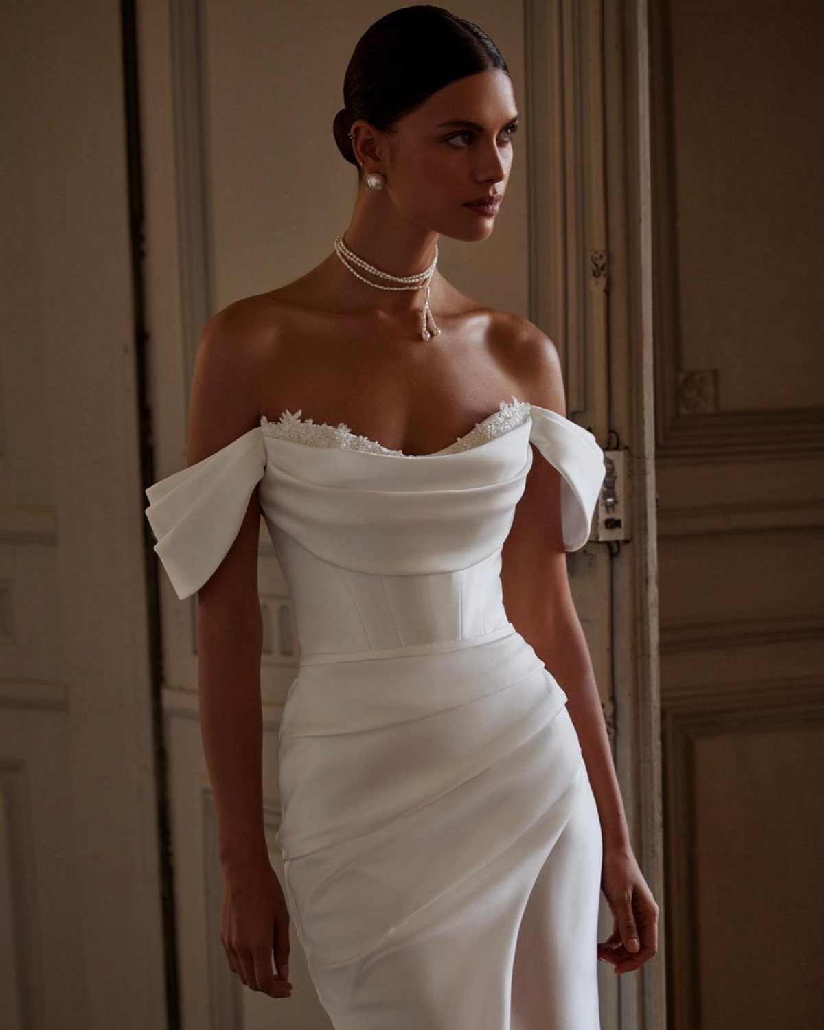 Woman posing in a white off-the-shoulder satin wedding dress with draped details.