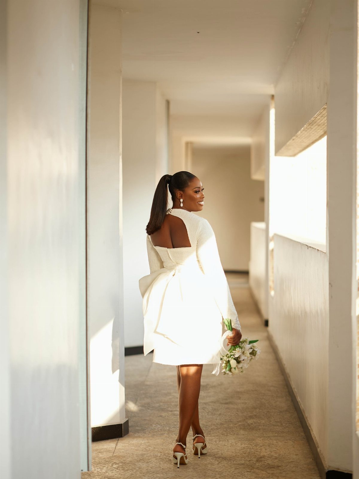 A woman in a short white minimalist wedding dress walks through a bright hallway holding flowers.