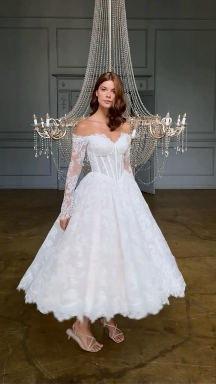 Woman in a white lace off-the-shoulder tea-length wedding dress standing before a chandelier.