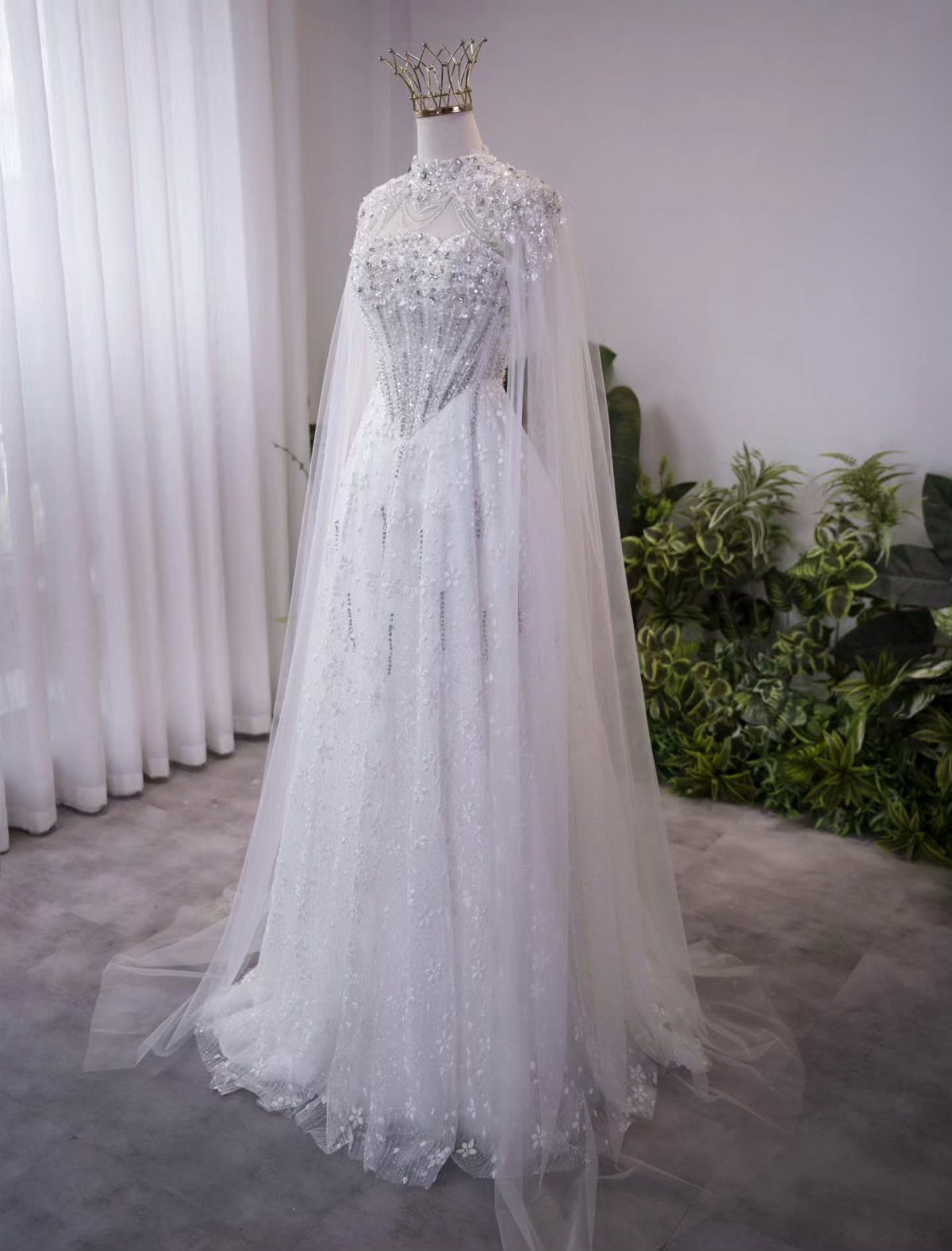 A sparkling white wedding dress with a sheer cape and a gold crown on a mannequin.