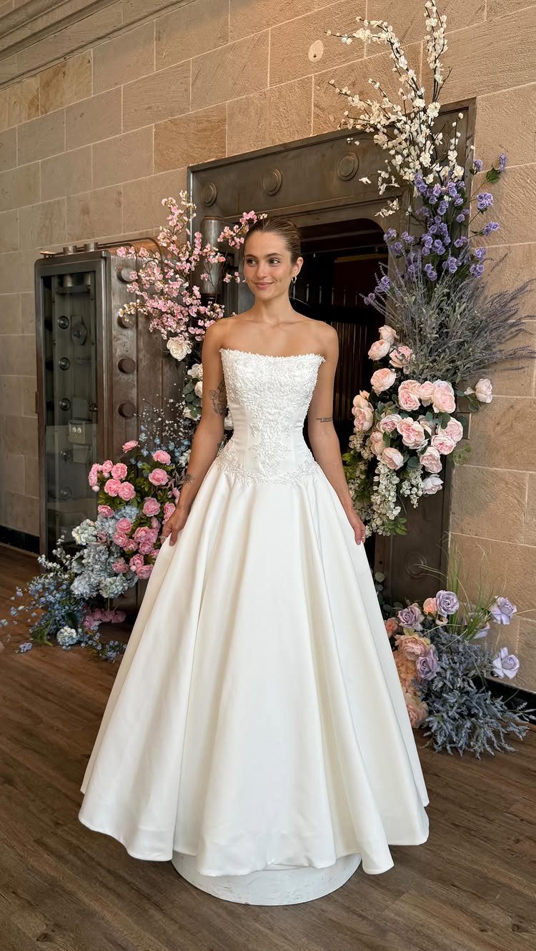 Woman in a strapless white wedding dress standing amidst large pink and blue floral arrangements.