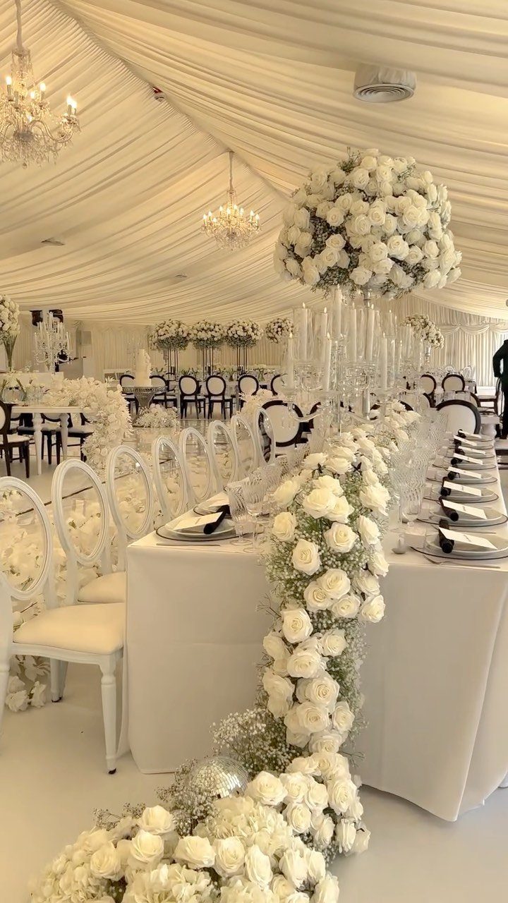 White rose garlands and floral centerpieces decorate an elegant banquet table with white linens and crystal chandeliers.