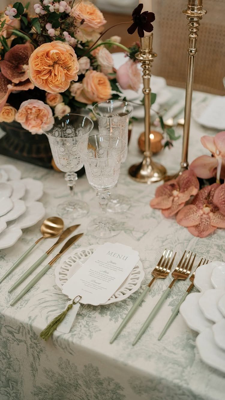 Elegant table setting with patterned tablecloth, gold cutlery, crystal glasses, and peach floral arrangements.