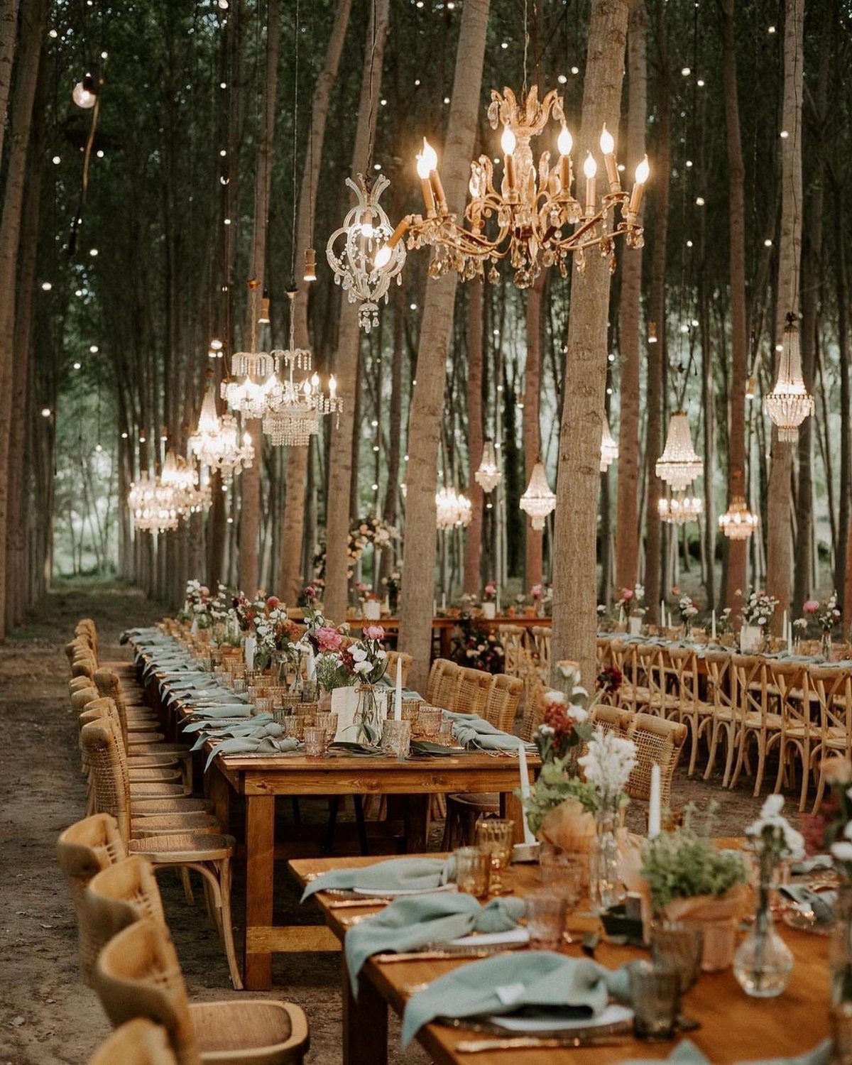 Enchanted Forest Wedding Theme: 10 Ideas for a Natural Setting 8 Long wooden dining tables set with flowers and glassware under hanging crystal chandeliers in a forest.