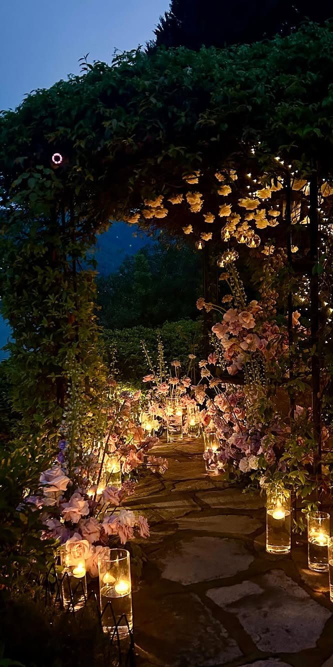Glowing glass cylinder candles line a stone pathway surrounded by pink flowers and greenery.