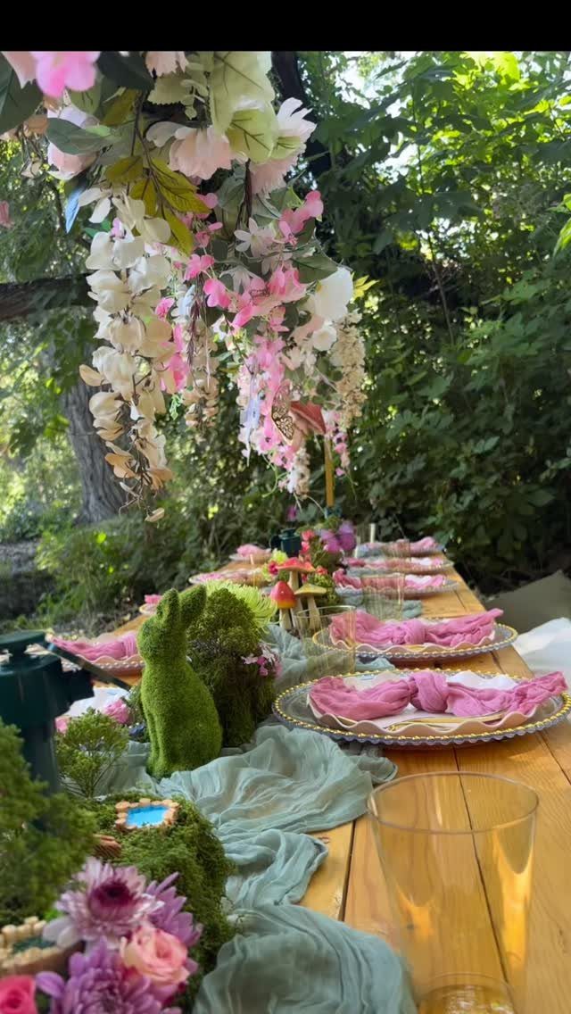 A wooden table decorated with moss, pink flowers, and sage green fabric under hanging blossoms.