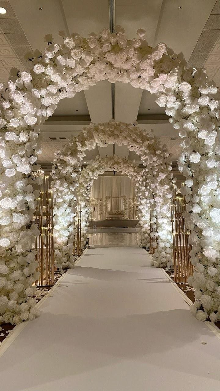 White floral arches forming a tunnel over a white aisle runner in an indoor venue.