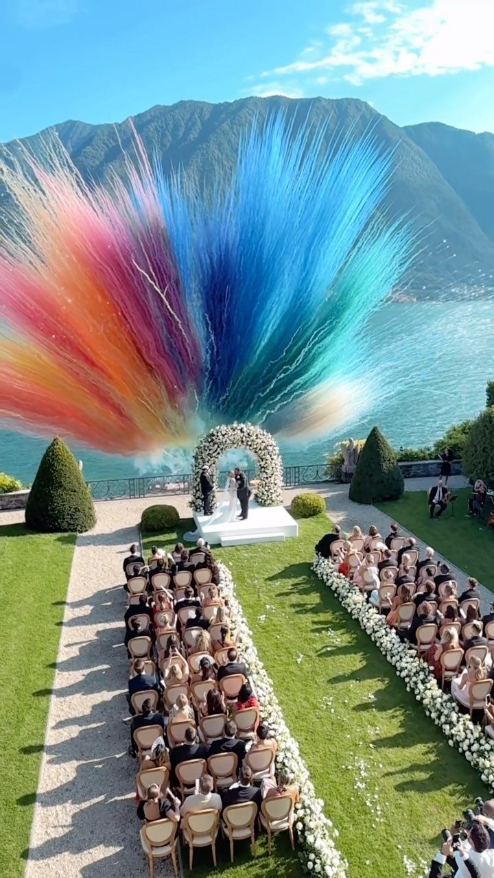 Wedding guests seated on a lawn facing a floral arch overlooking a lake and mountains.