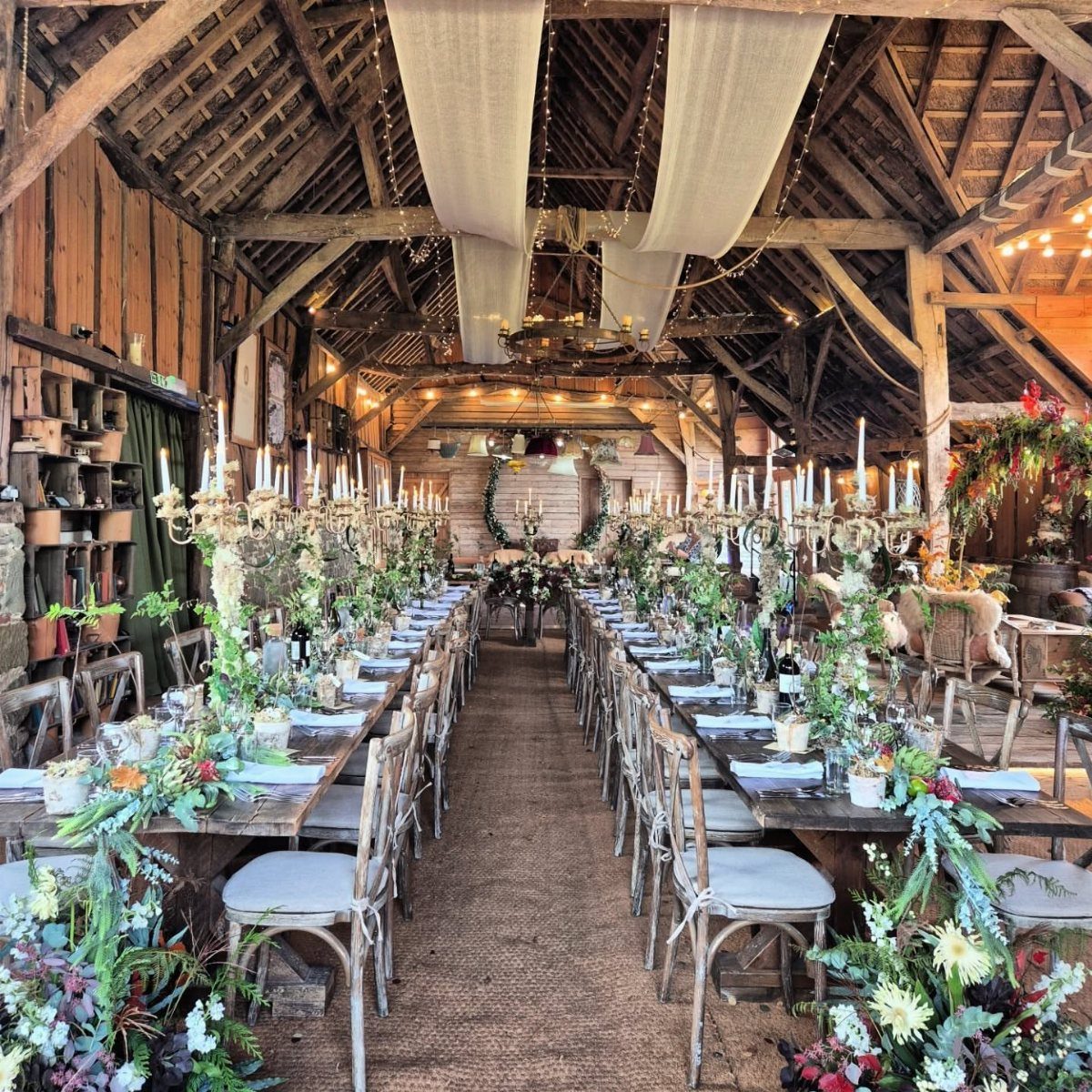 Long wooden banquet tables decorated with greenery and candles inside a rustic timber barn.