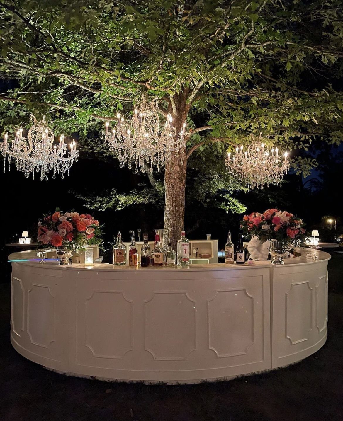 A white curved bar decorated with flowers and liquor under hanging crystal chandeliers.