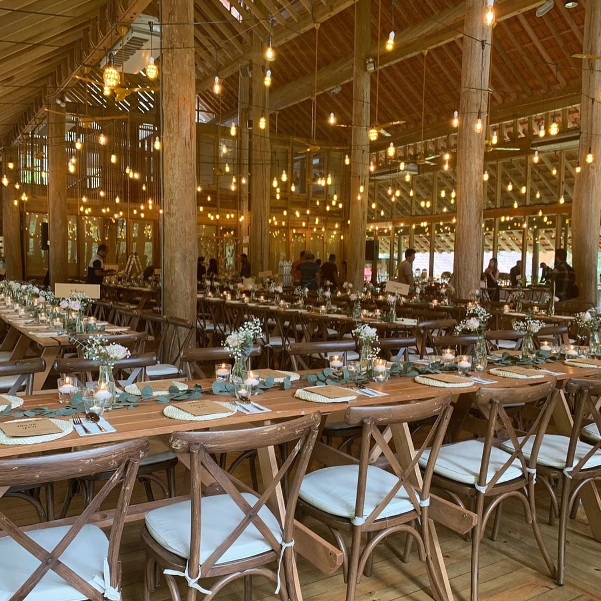 Long wooden banquet tables set with greenery and candles inside a rustic wooden pavilion.