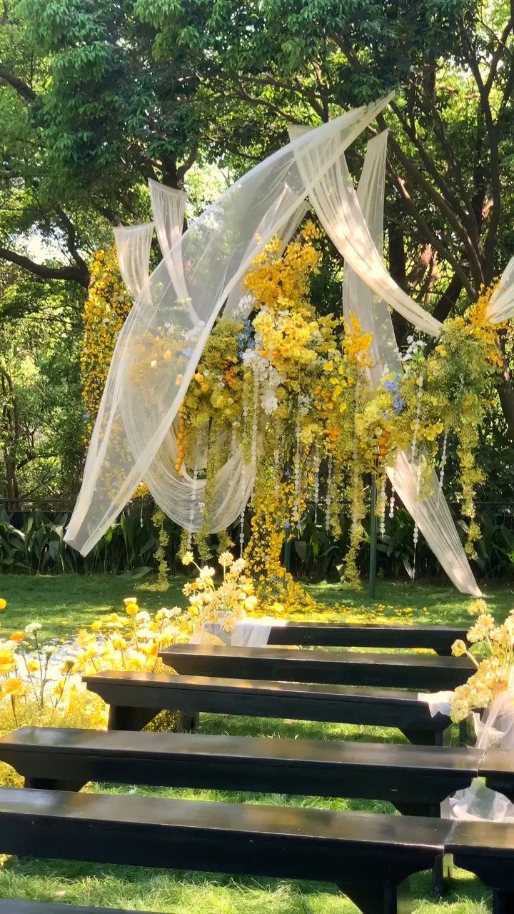 Fall Wedding Color Palette: 10 Ideas Photographed on Real Tables 5 Yellow floral wedding arch with white fabric draping over dark wooden benches outdoors.