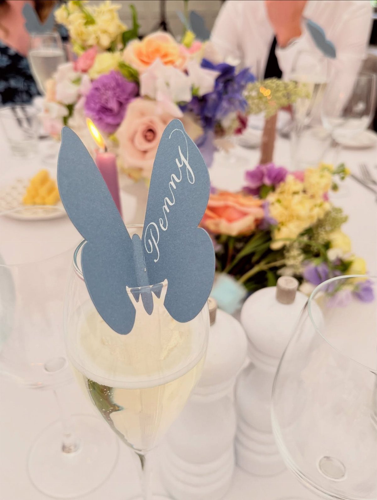 A blue butterfly-shaped place card with the name Penny sits on a champagne glass.