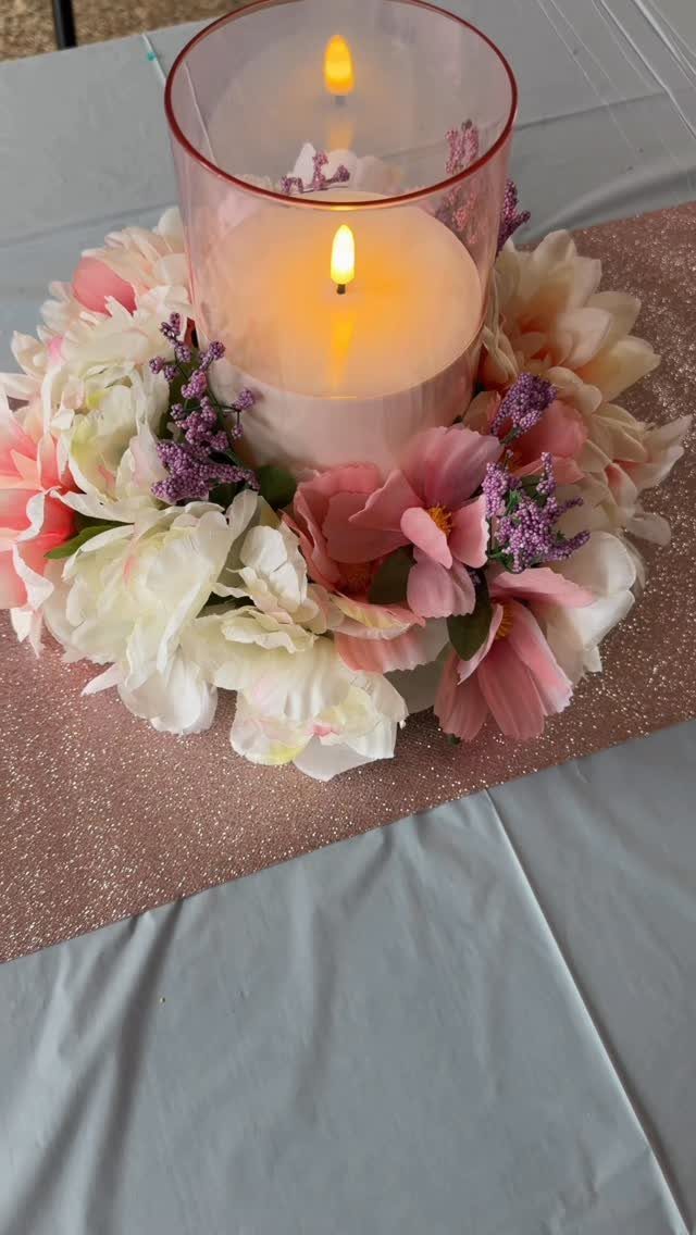 A white candle in a glass cylinder surrounded by cream and pink flowers on a glittery runner.