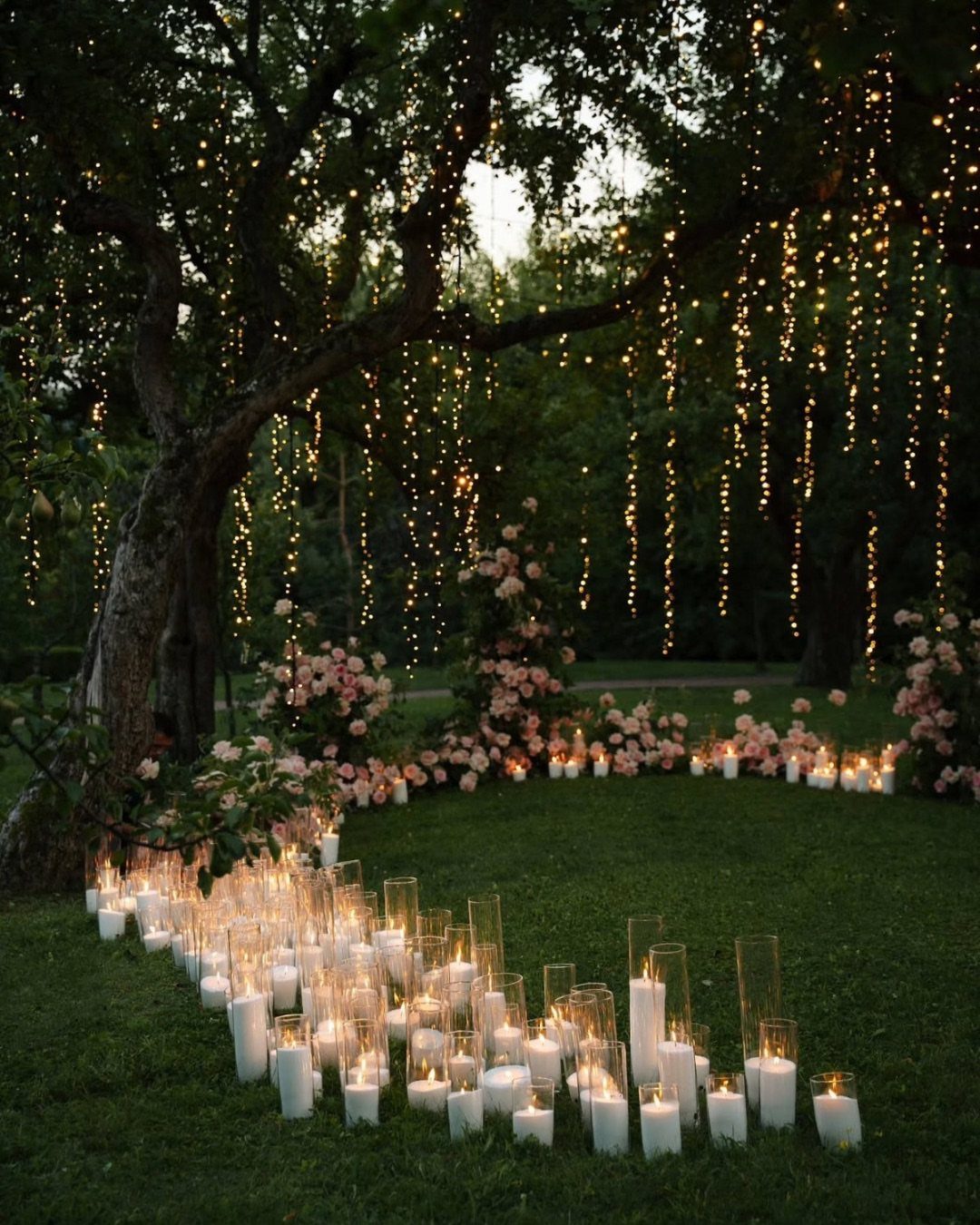 Hanging fairy lights on a large tree and lit candles in glass vases on a garden lawn.