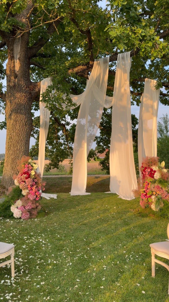 White fabric drapes hanging from a large tree surrounded by pink floral arrangements in a garden.