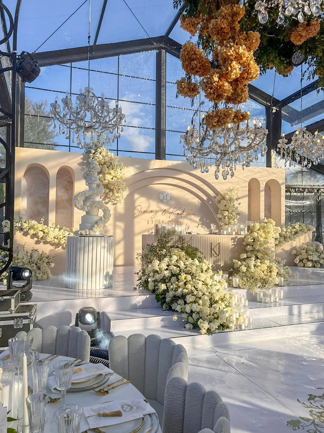 Elegant wedding reception setup with white flowers, crystal chandeliers, and gold cutlery on white tables.