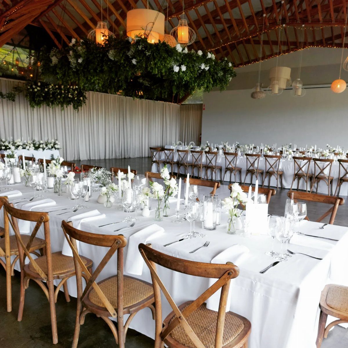 Long white dining tables set with flowers under large hanging greenery and warm lanterns.
