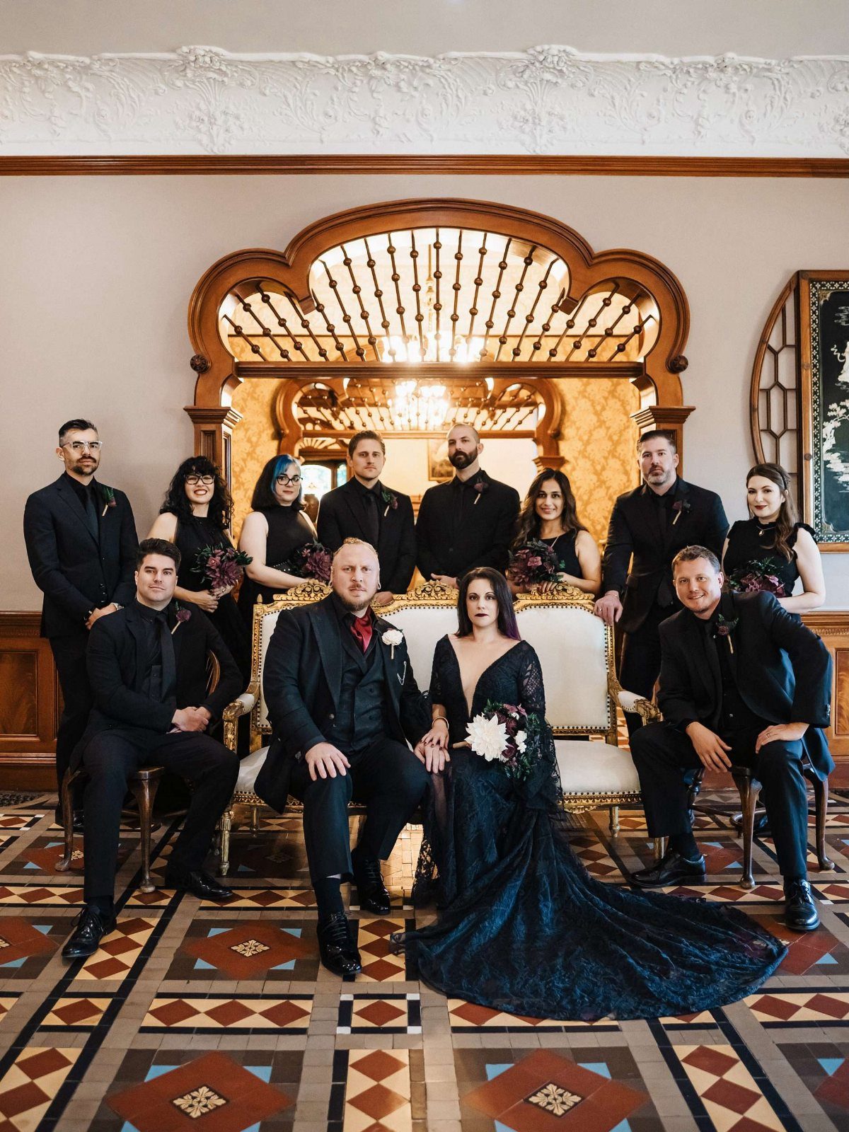 Gothic Wedding Theme: 11 Dark & Elegant Ideas 10 A wedding party dressed in black attire posing in an ornate room with patterned flooring.