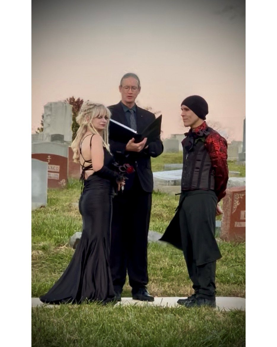 Gothic Wedding Theme: 11 Dark & Elegant Ideas 9 A bride and groom stand before an officiant during a gothic wedding ceremony in a cemetery.