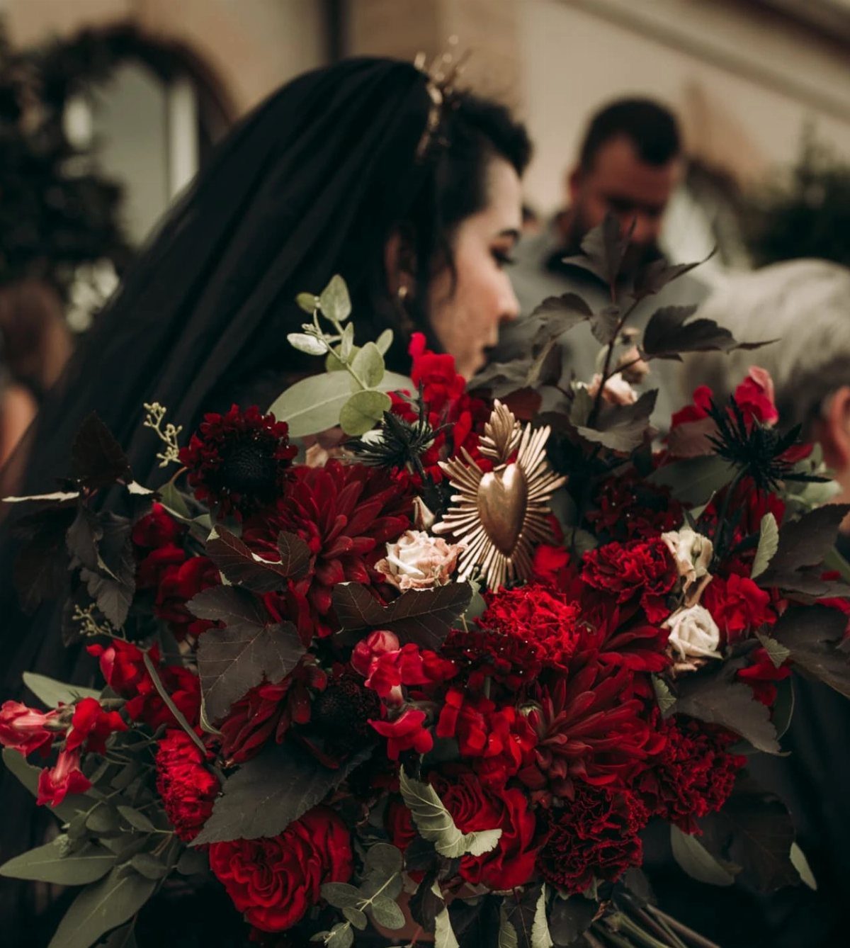 Gothic Wedding Theme: 11 Dark & Elegant Ideas 5 A dark bridal bouquet featuring deep red roses, burgundy dahlias, and a gold heart ornament.