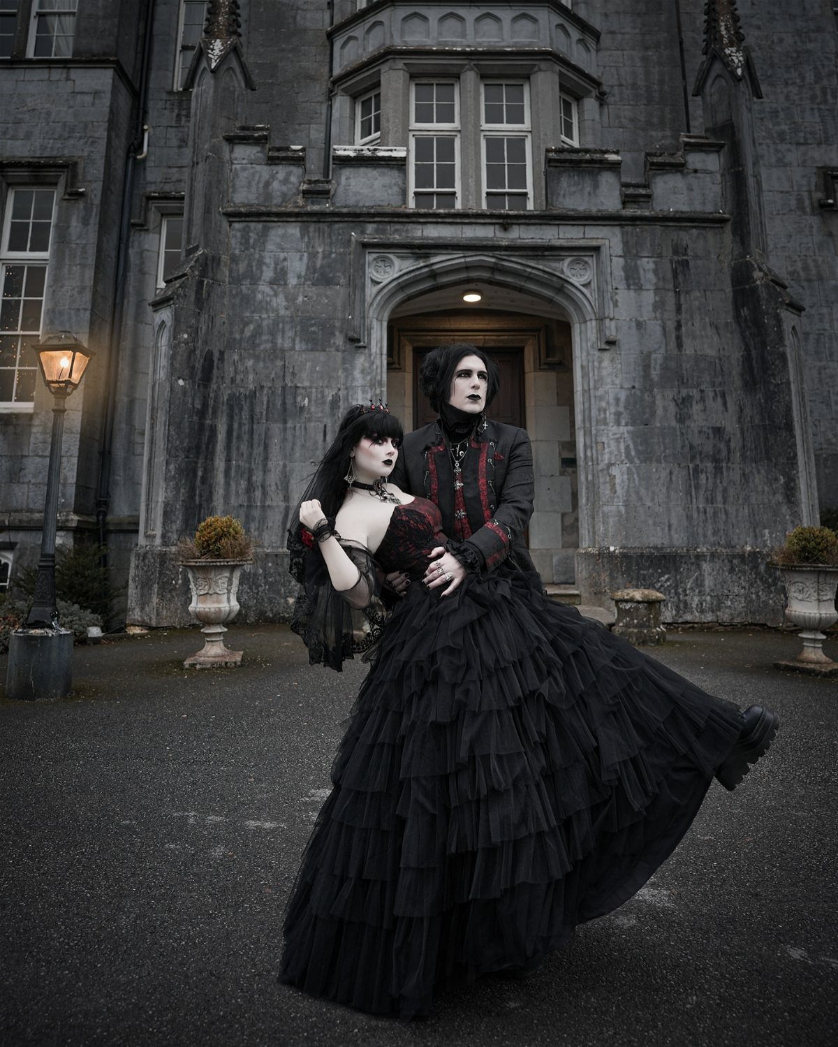 Gothic Wedding Theme: 11 Dark & Elegant Ideas 2 A couple wearing dramatic black gothic wedding outfits posing before a large stone castle.