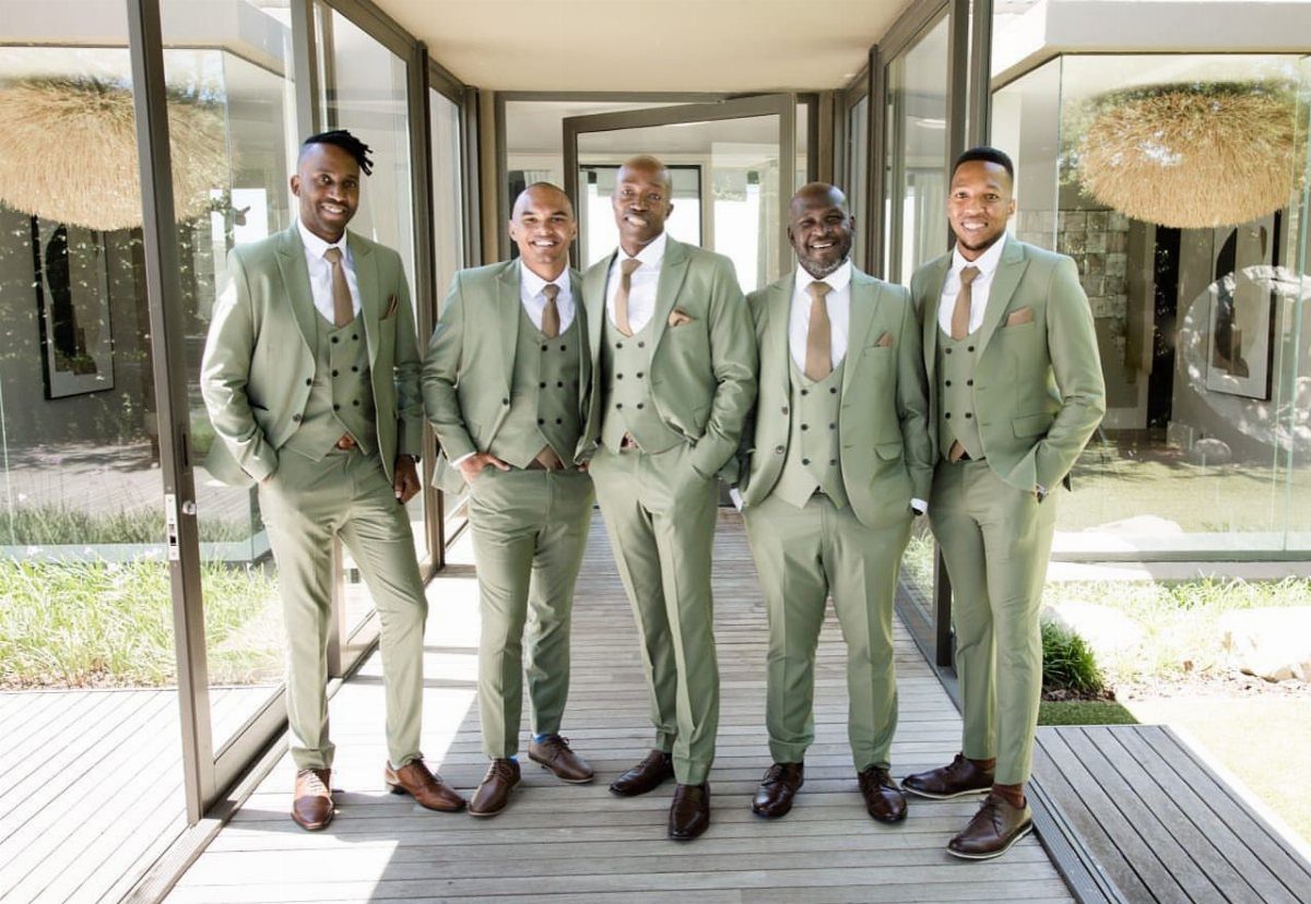 Green Wedding Suit: 8 Looks for Grooms and Guests 2 Five men posing in matching sage green three-piece suits on a wooden walkway.