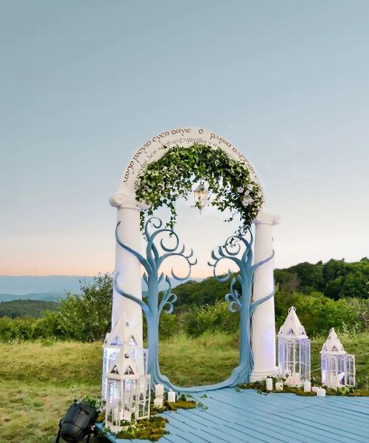 Hobbit Themed Wedding: 20 Ideas for Your Shire Celebration 17 A white wedding arch with blue tree designs and Elvish script overlooking a green landscape at sunset.
