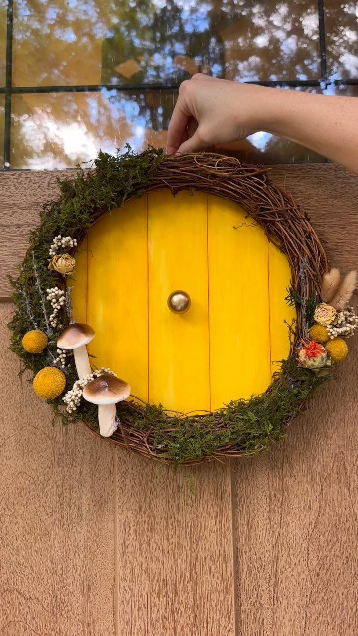 Hobbit Themed Wedding: 20 Ideas for Your Shire Celebration 3 A hand holds a rustic wreath made of moss, twigs, mushrooms, and yellow flowers against a yellow door.