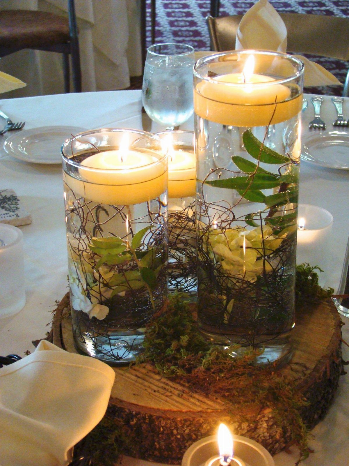 Lord of the Rings Centerpieces: 7 Tabletop Ideas for Your Event 6 Floating cream candles in tall glass cylinders with green leaves on a mossy wood slice.