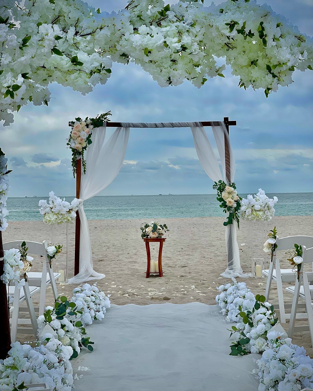 16 Marriage Theme Ideas for 2026: From Beach Ceremonies to Pearl Aesthetics 7 A white floral wedding ceremony setup on a sandy beach overlooking the ocean.