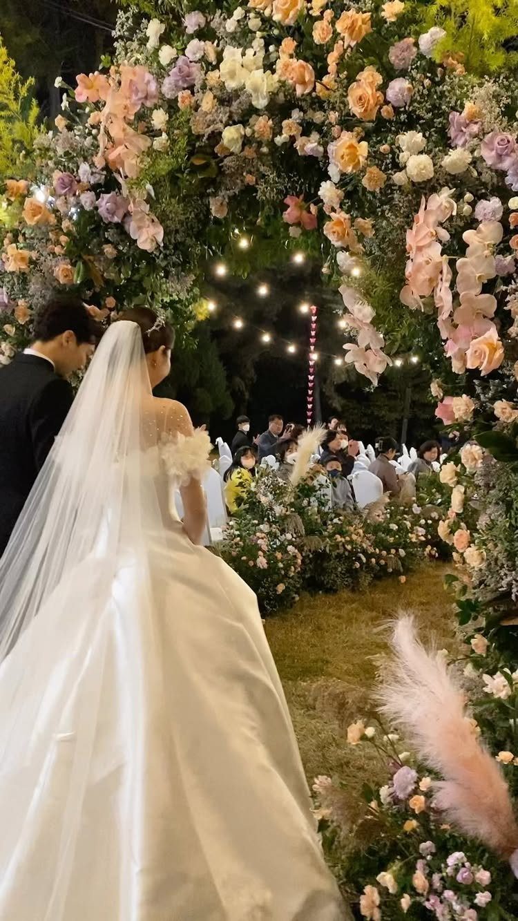 16 Marriage Theme Ideas for 2026: From Beach Ceremonies to Pearl Aesthetics 2 Bride and groom standing before a large archway of pastel pink and peach flowers.