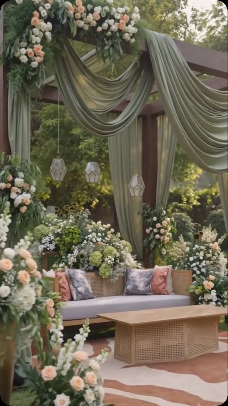 16 Marriage Theme Ideas for 2026: From Beach Ceremonies to Pearl Aesthetics 9 Garden lounge with green draped fabric, floral arrangements, a sofa, and a rattan coffee table.