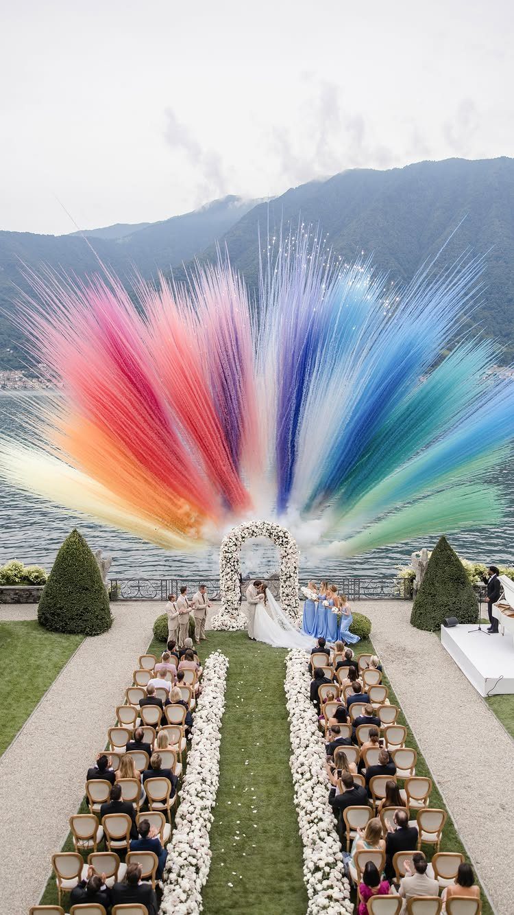 16 Marriage Theme Ideas for 2026: From Beach Ceremonies to Pearl Aesthetics 5 A wedding ceremony with a massive rainbow-colored spray effect behind a floral arch by a lake.